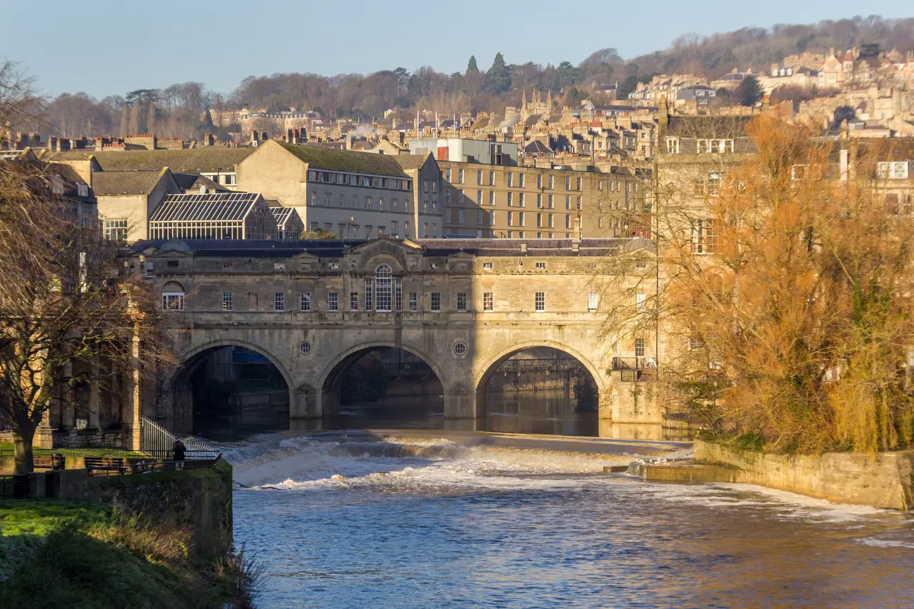 Pulteney Bridge in Bath with its three arches above the curved weir on the River Avon, autumn trees beside it and Georgian terraces on the hill behind