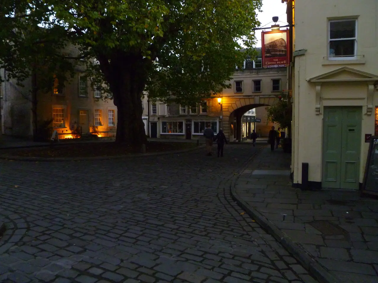 Cobbled Abbey Green in Bath at dusk under a big plane tree, with the Crystal Palace pub sign on the right and people walking towards an arch