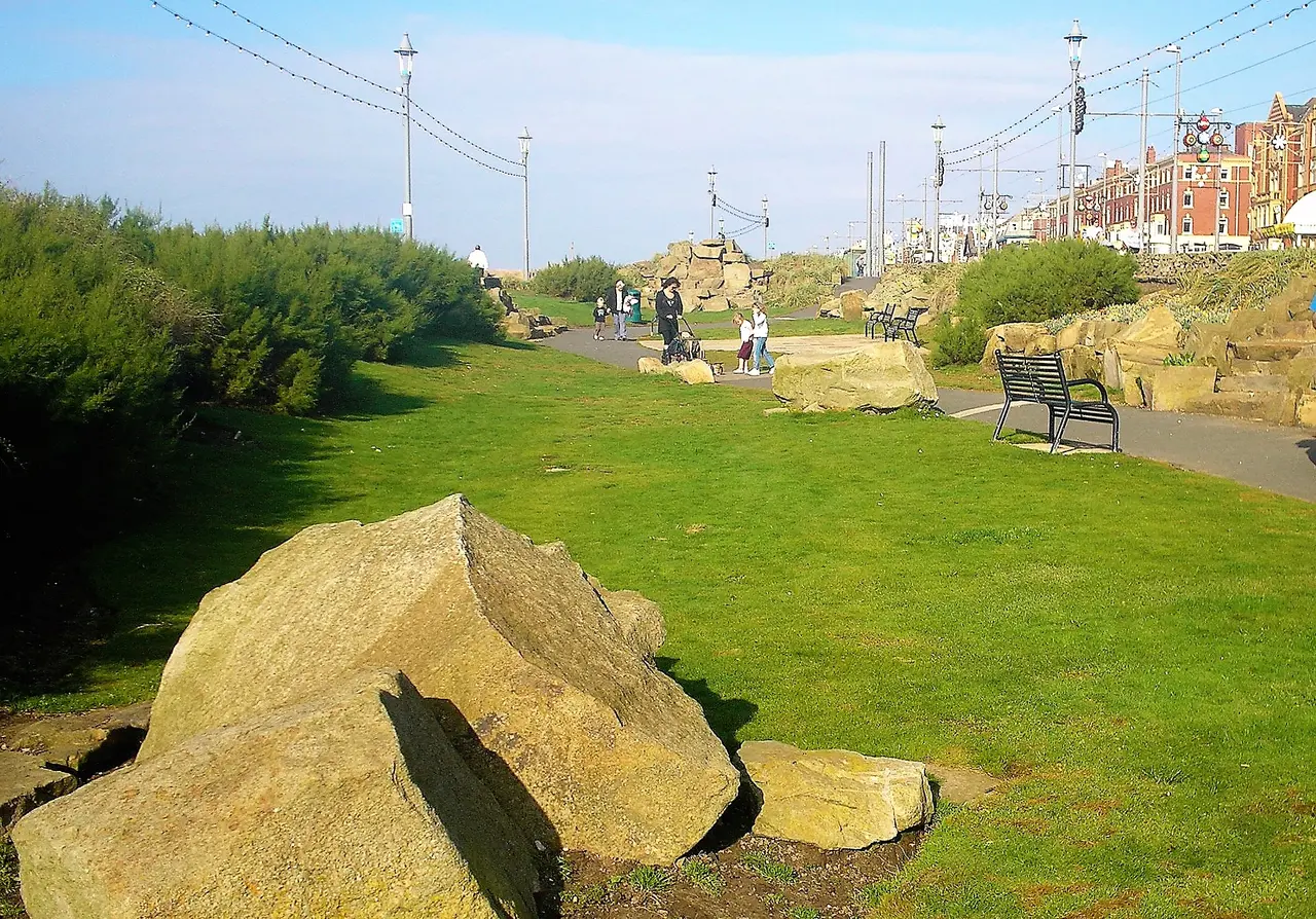 Grassy cliff gardens on North Shore with large sandstone rocks, a bench and people walking along the path towards the hotels on the promenade