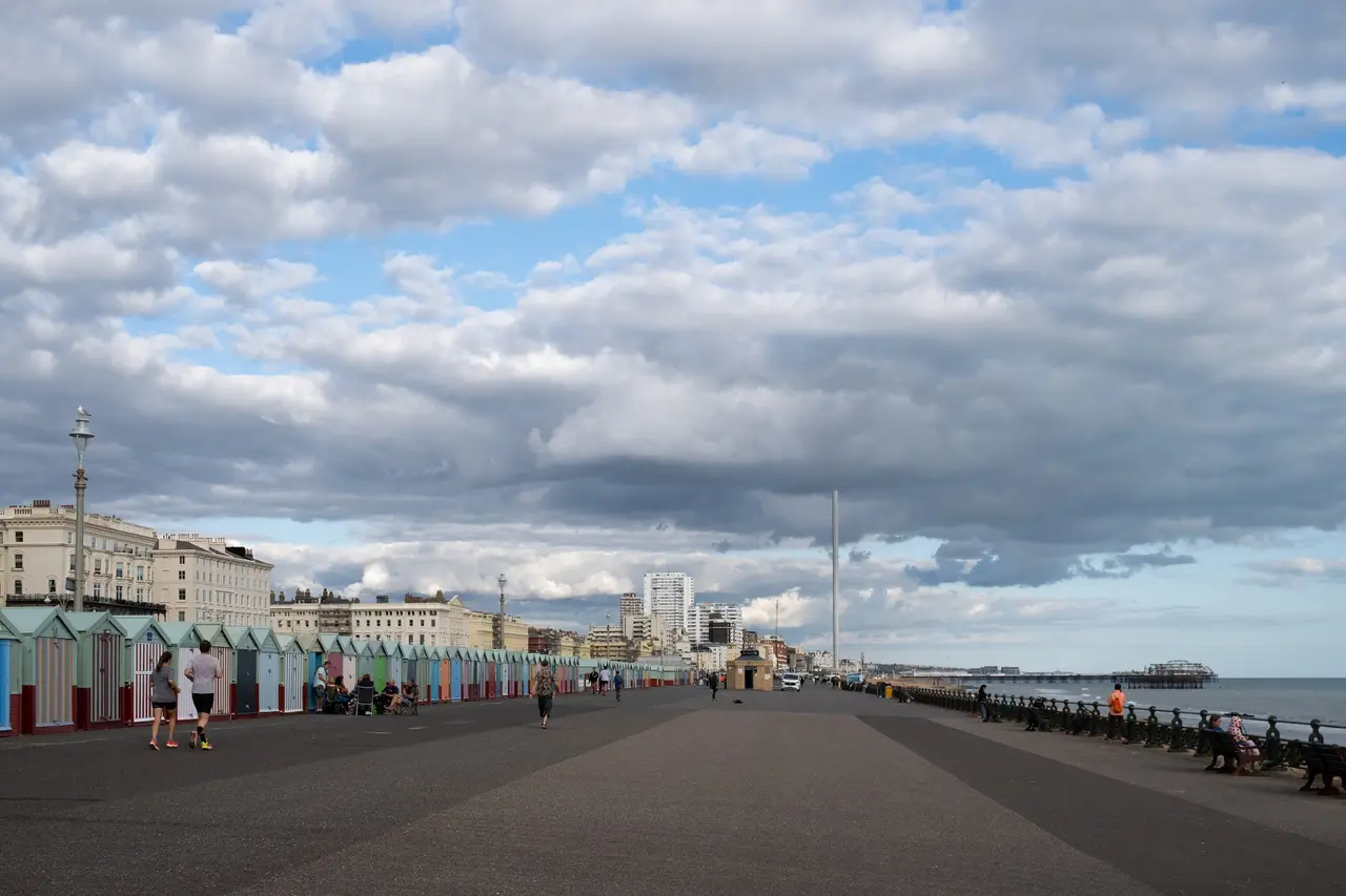 Hove promenade on a cloudy day, with a row of coloured beach huts on the left, people walking, and the i360 tower and seafront hotels in the distance