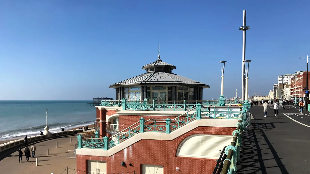Brighton's seafront bandstand with its green railings and domed roof, on the promenade above the beach, with the i360 tower behind under a clear blue sky