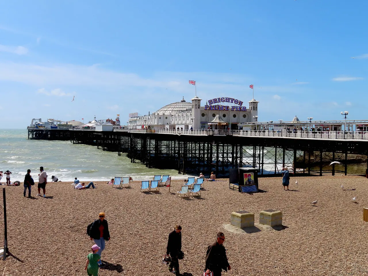 Brighton Palace Pier on a sunny day, with people walking on the pebbles and striped deckchairs on the beach in front