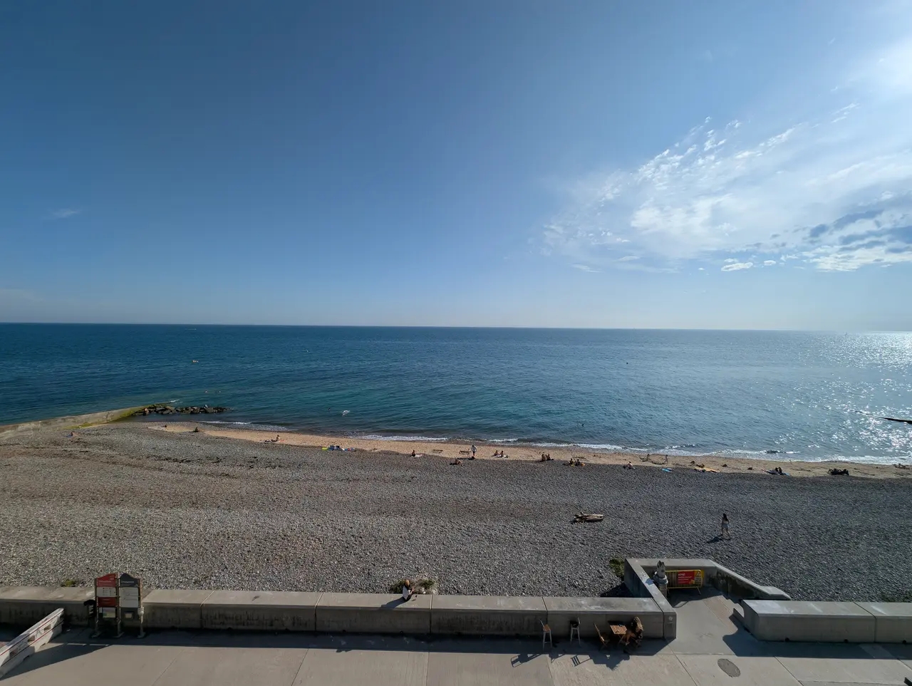Looking down on a long shingle beach and the calm blue sea from the clifftop at Ovingdean, with a few people on the sand and the Undercliff Walk below