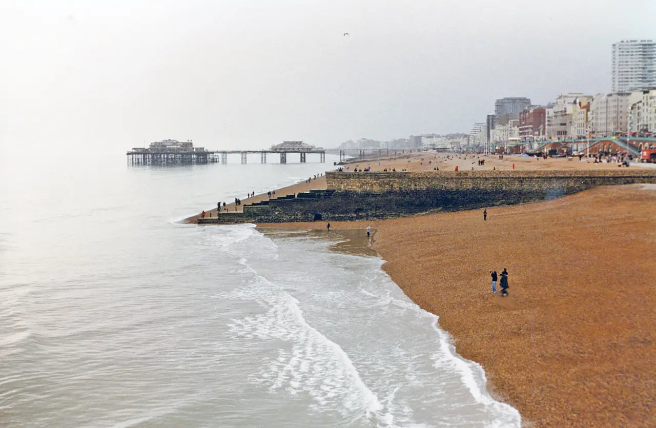 A wide shingle beach in Brighton with gentle waves, a couple walking at the water's edge and the West Pier and seafront buildings in the distance