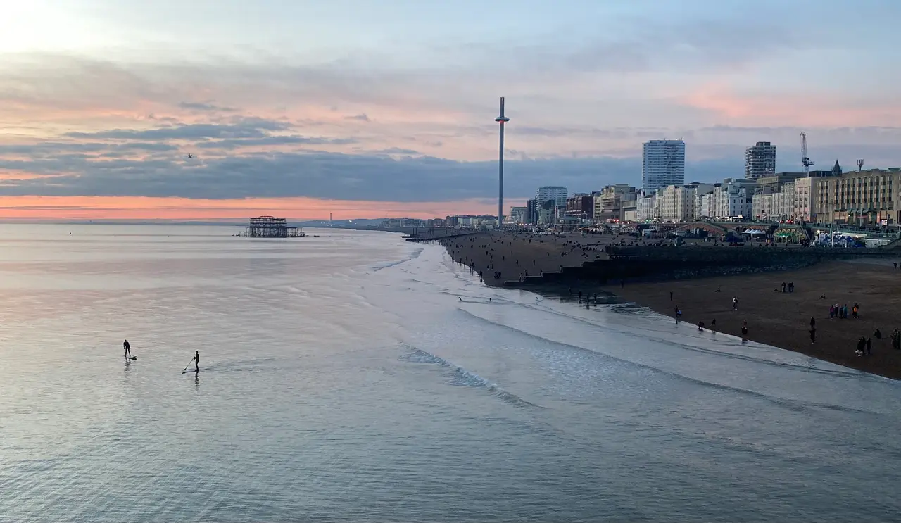 Brighton's seafront at dusk seen from the pier, with the shingle beach curving away, the ruined West Pier and the i360 tower against a pink sky, and two paddleboarders on the calm sea