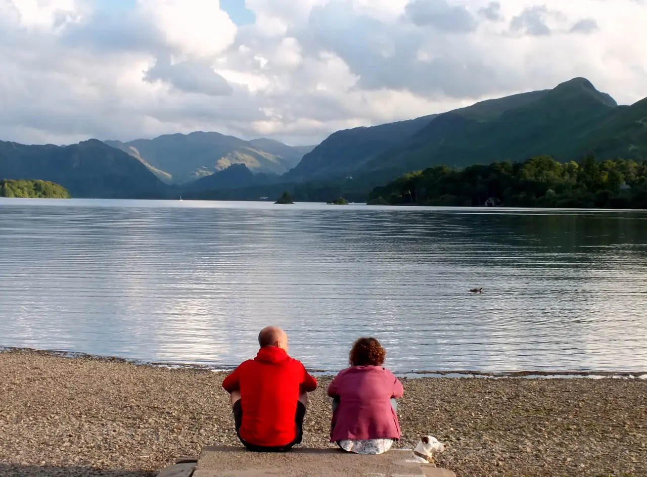 Two people sitting on the shingle shore of Derwent Water with a small white dog beside them, looking across the calm lake to Borrowdale's fells
