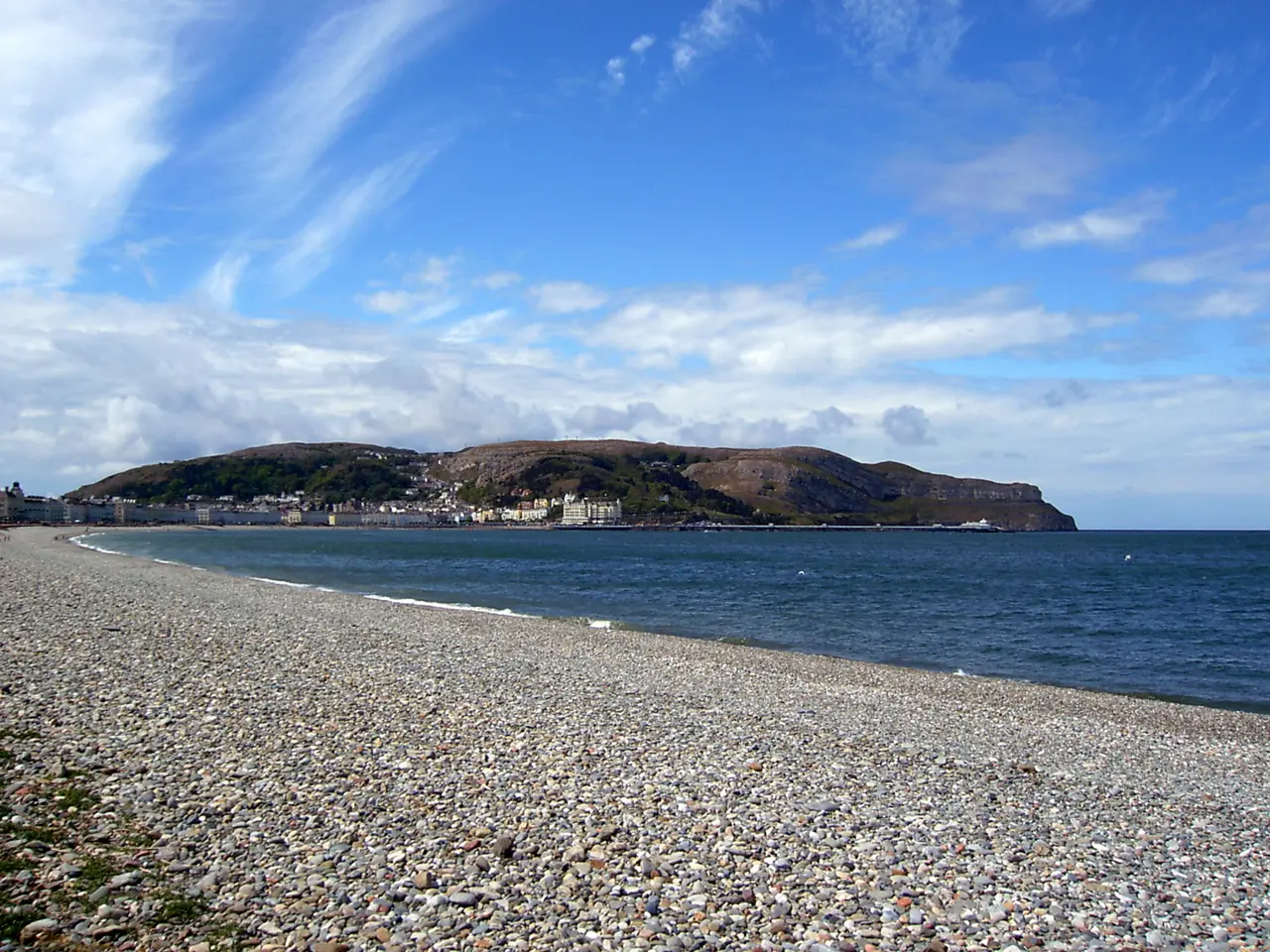 The Great Orme seen across the bay from the pebbles at the Craig-y-Don end of the North Shore, under a blue sky with high cloud