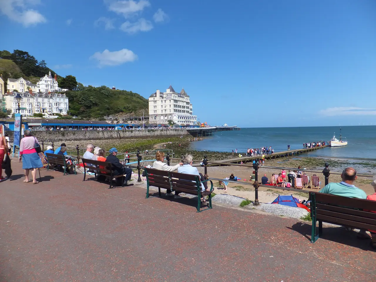 People on benches along the promenade on a sunny day, looking across the North Shore to the Grand Hotel, the pier and the Great Orme