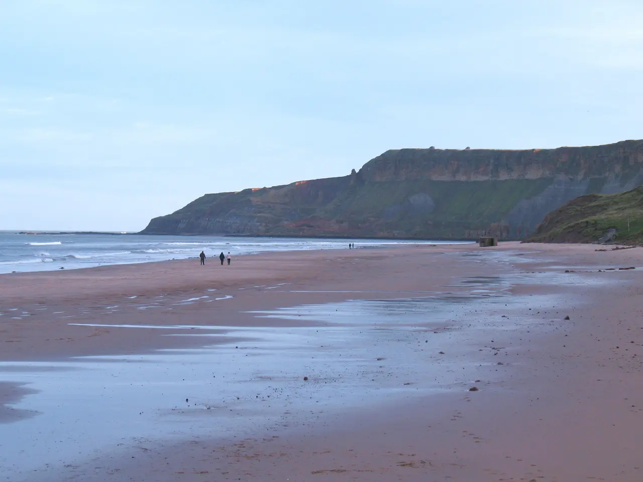 Wide wet sands at Cayton Bay at low tide, with a few walkers near the water and a dark headland at the end of the bay