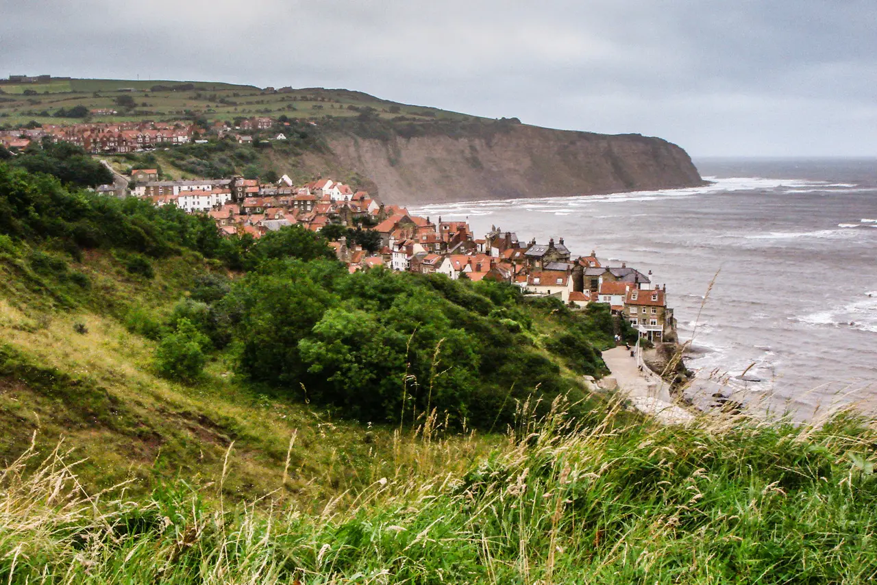 Robin Hood's Bay seen from the clifftop path: red-roofed houses tumbling down to the sea, with the headland of Ravenscar beyond