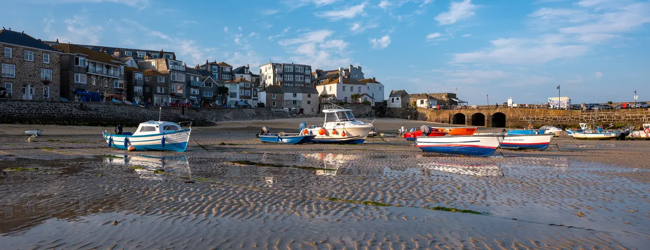 Small fishing boats sitting on the wet sand of St Ives harbour at low tide, below the stone houses along the Wharf, under a blue sky