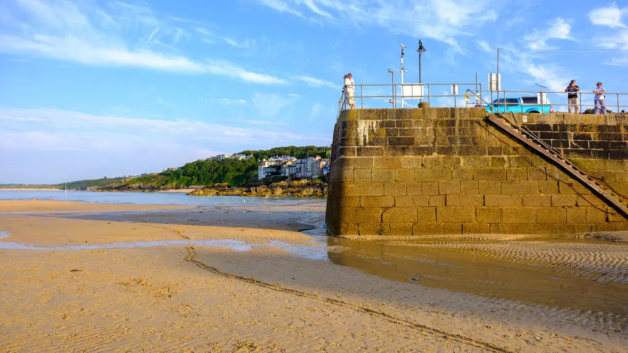 The granite wall of a pier in St Ives harbour rising from the sand at low tide, with people at the railings on top and a wooded headland beyond
