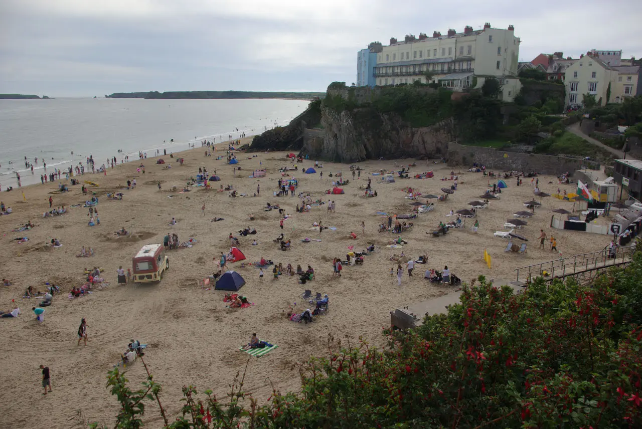 Castle Beach seen from above on a busy summer day, with a cream hotel terrace on the cliff and red flowers in the foreground