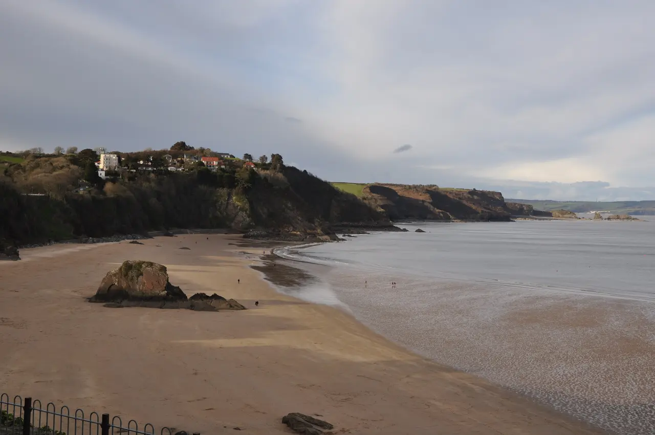 Goskar Rock standing on the empty sand of North Beach at low tide, below wooded cliffs under a grey sky
