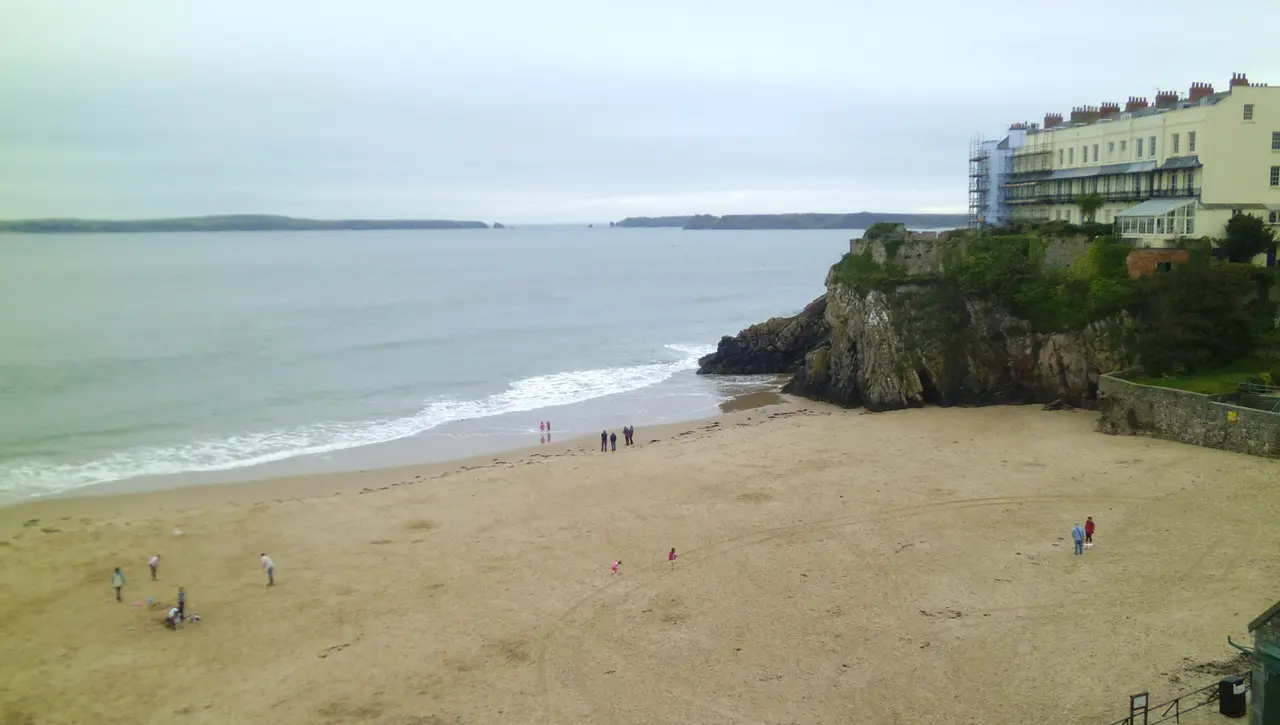 South Beach with waves breaking on wide sand, a rocky headland with a hotel terrace above it and Caldey Island across the water