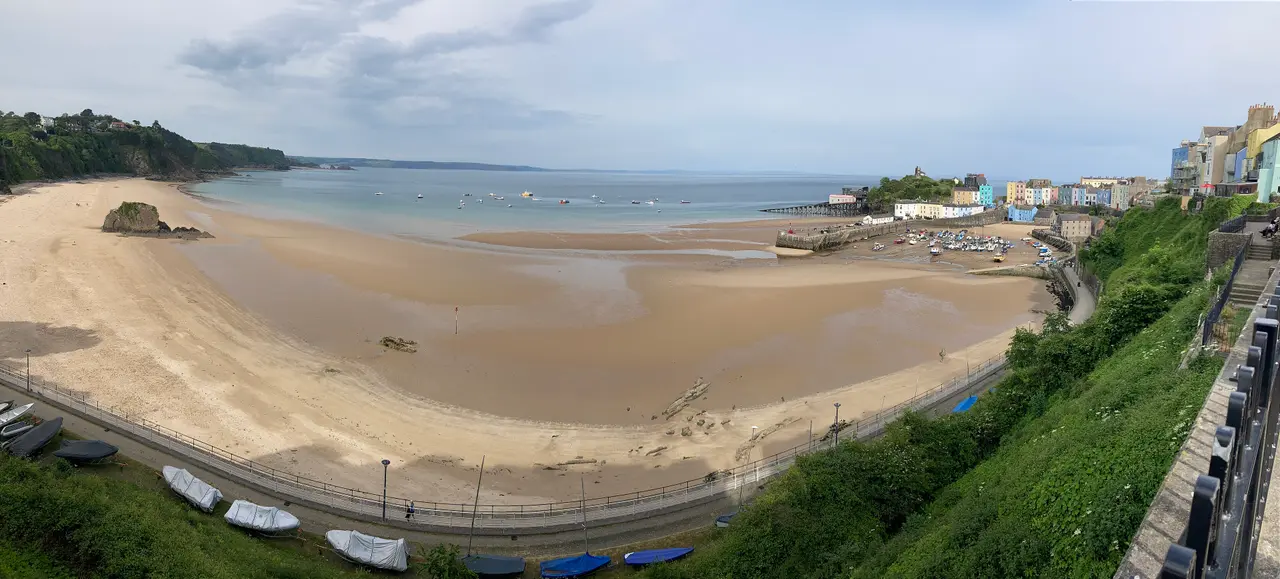 North Beach curving below a green slope, with Goskar Rock on the sand and the harbour and colourful houses of Tenby beyond, under a blue sky