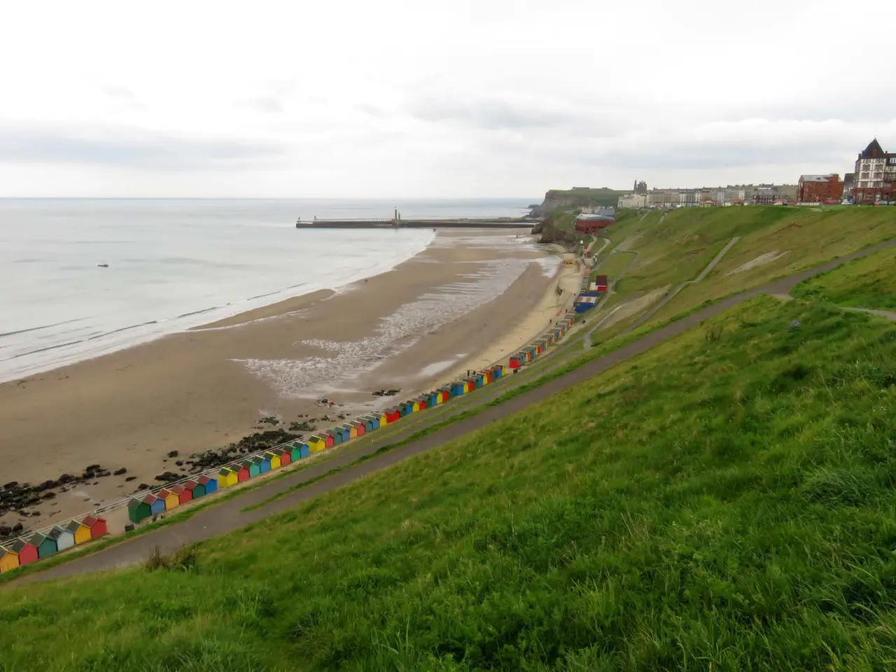 Whitby West Cliff beach at low tide from the grassy cliff slope, with beach huts along the foot of the cliff and the piers in the distance