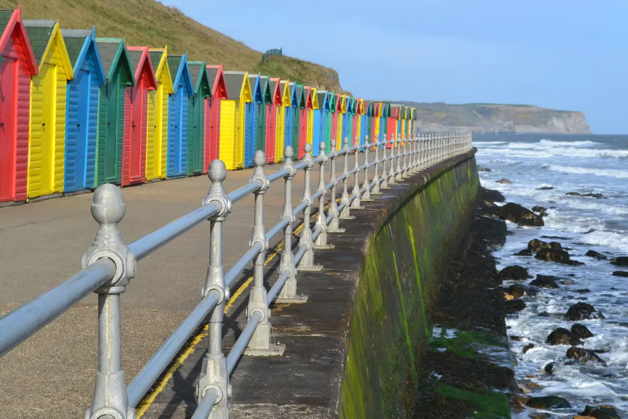 A long row of brightly painted beach huts on the promenade below Whitby's West Cliff, with waves breaking on the rocks beside it