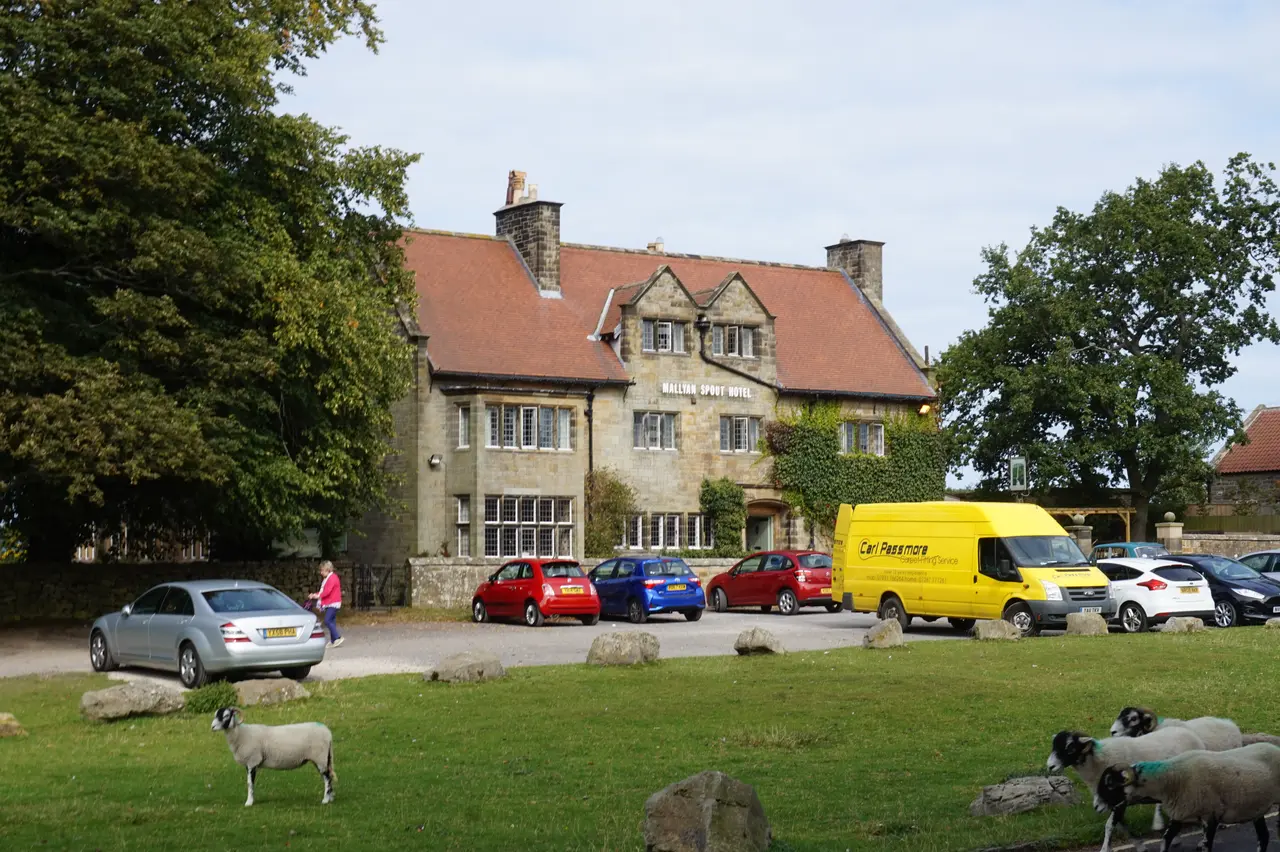 The stone-built Mallyan Spout Hotel behind Goathland's village green, with cars parked out front and sheep grazing on the grass